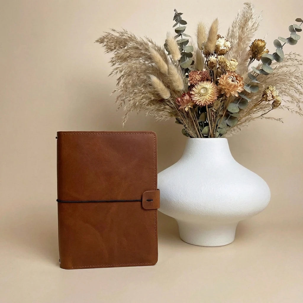 Brown leather notebook next to a white vase with dried flowers on a beige background