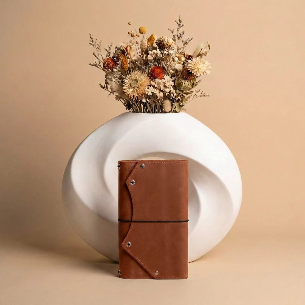 White vase with dried flowers and a brown leather notebook on a beige background