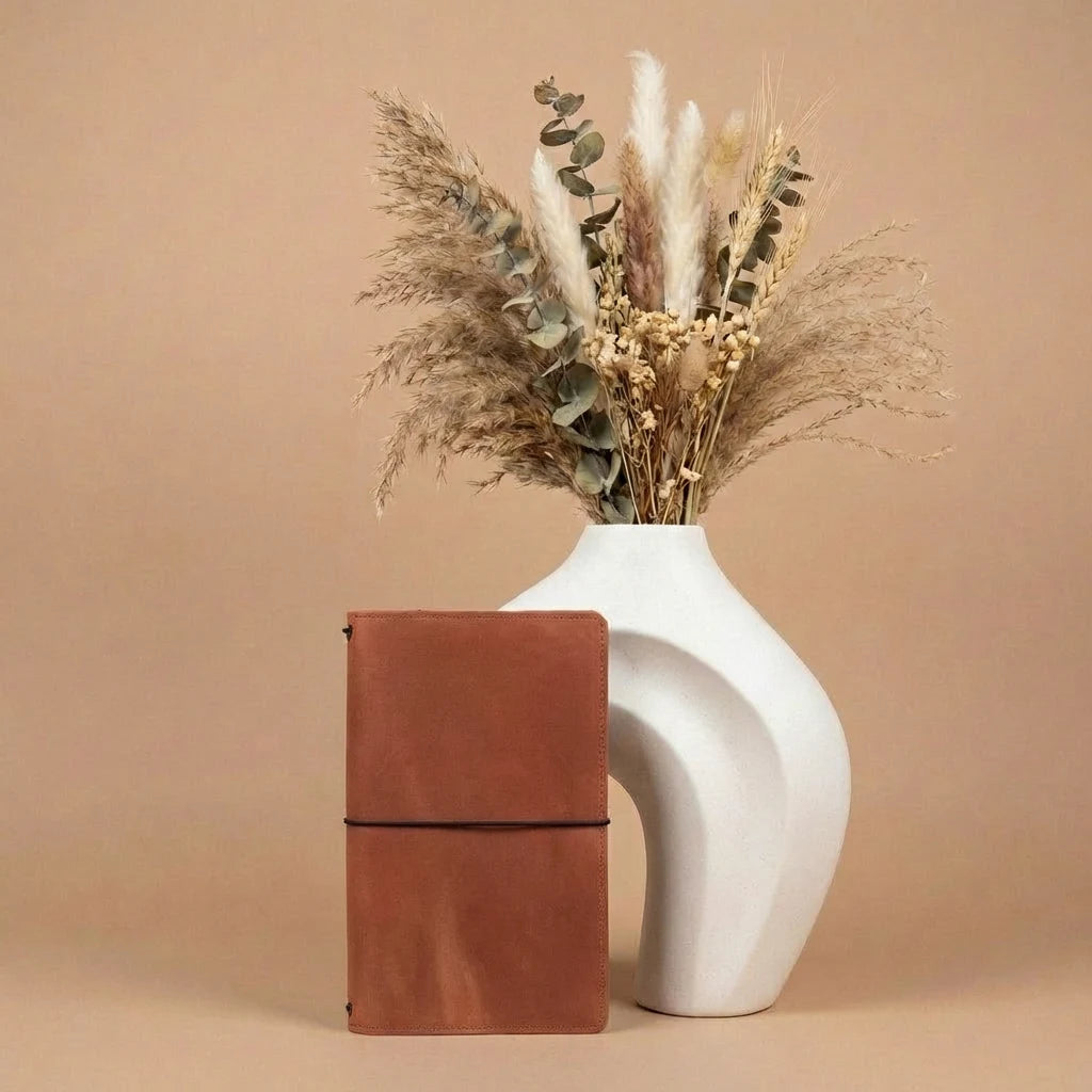 White vase with dried flowers and a brown leather notebook on a beige background