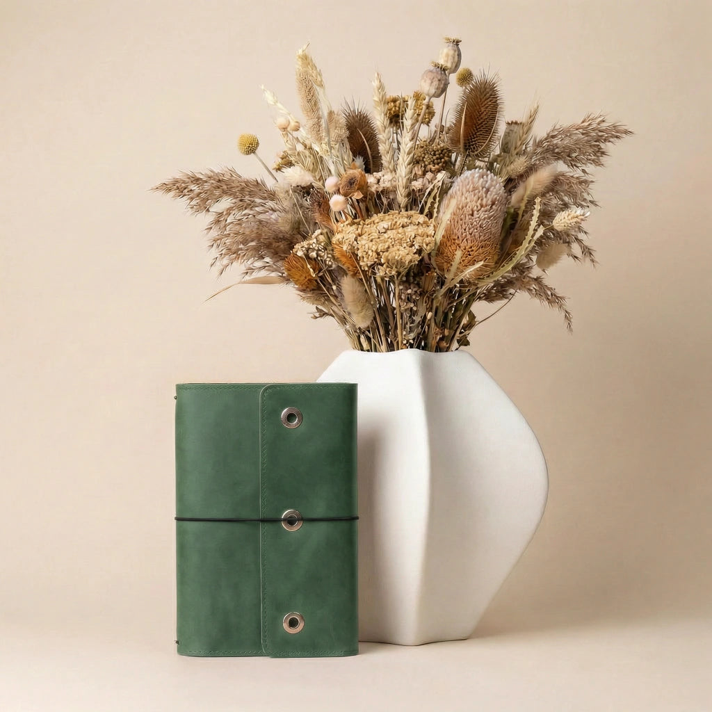 Green leather notebook next to a white vase with dried flowers on a beige background