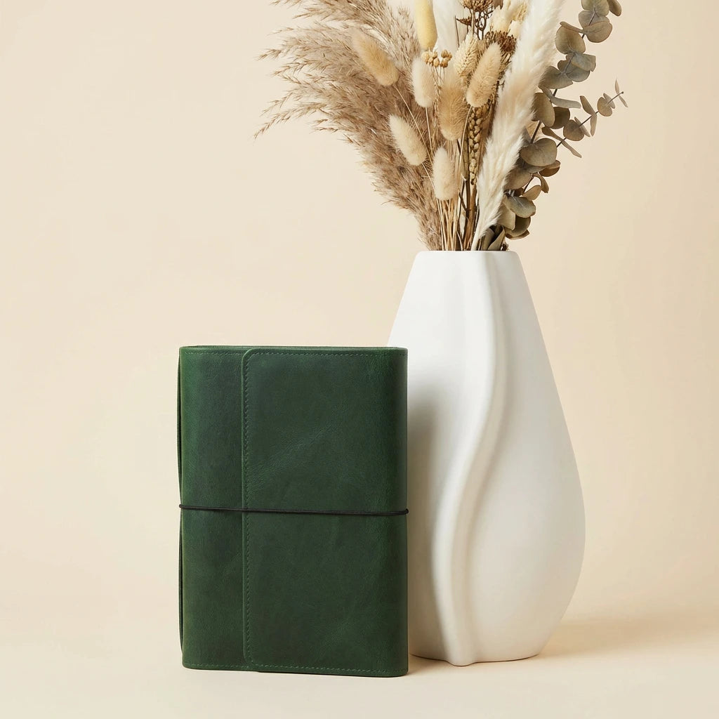 Green leather notebook next to a white vase with dried plants on a beige background