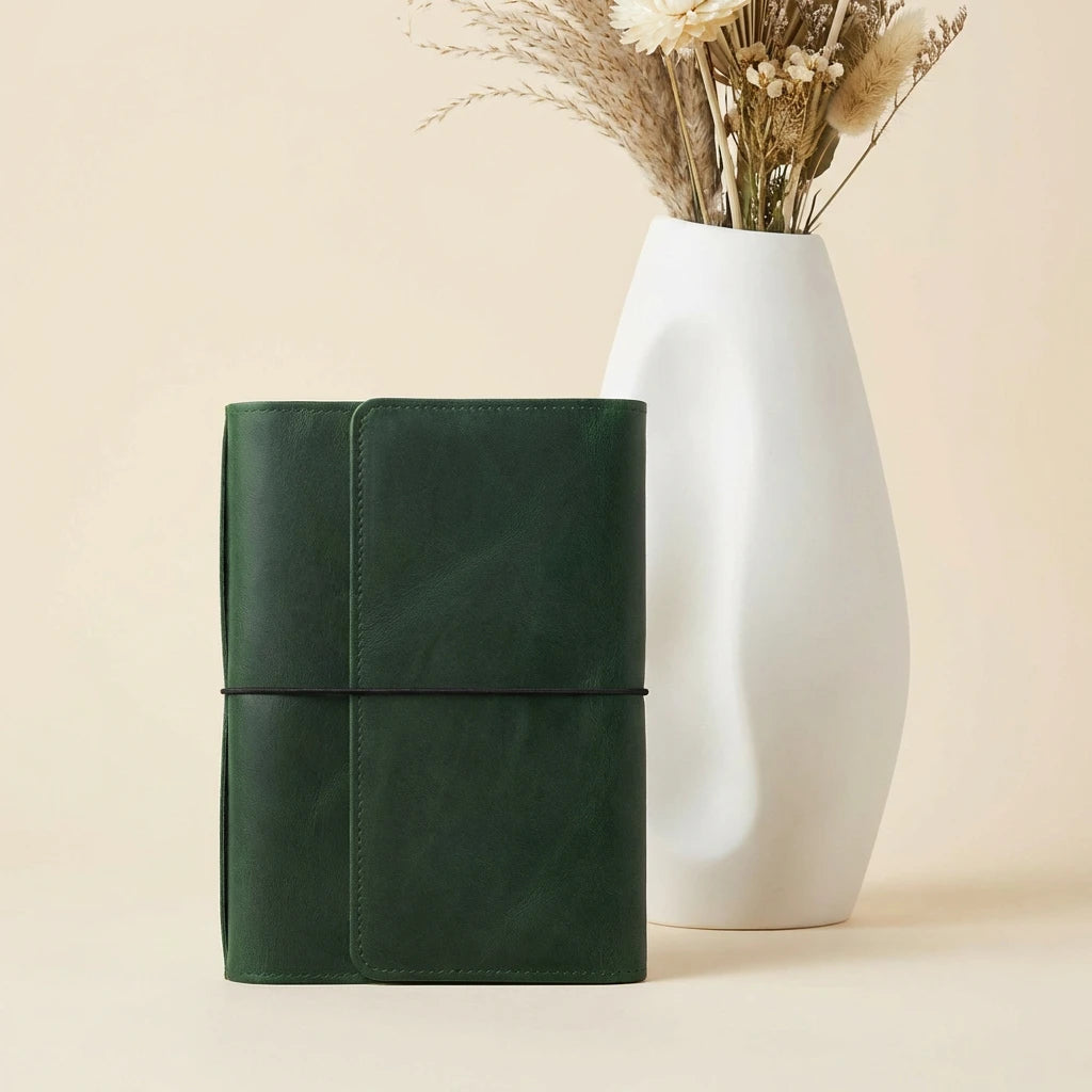 Green leather notebook next to a white vase with dried flowers on a beige background