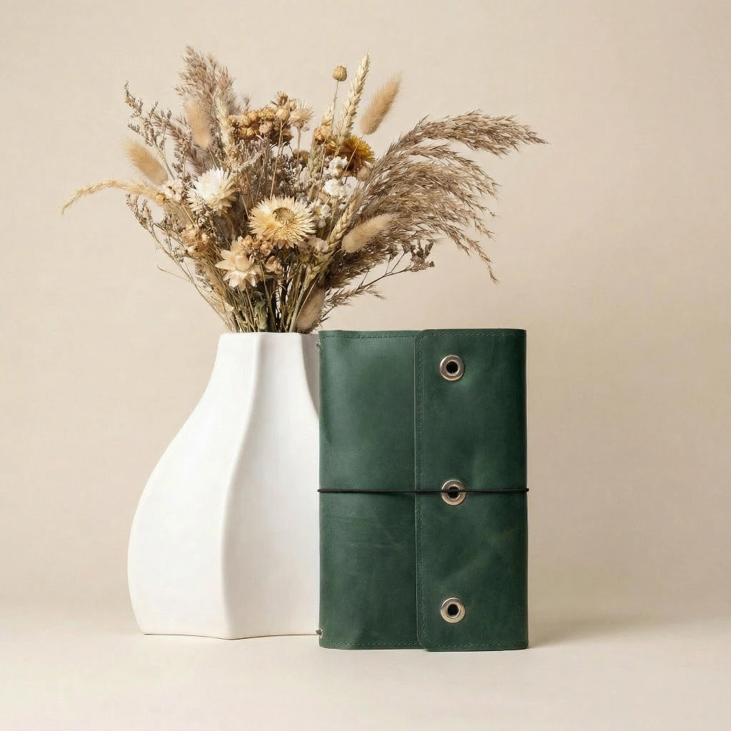 Green leather-bound notebook with a white vase containing dried flowers on a beige background