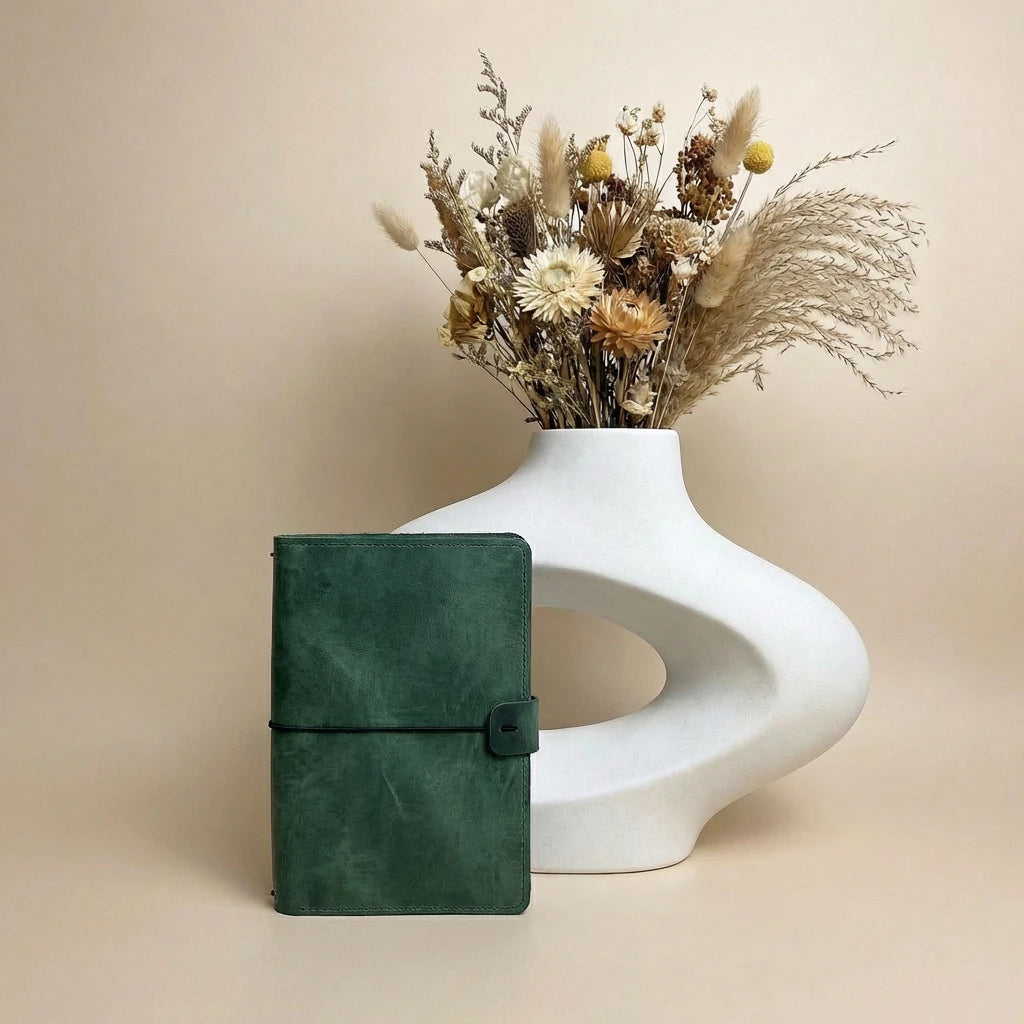 Green leather notebook next to a white vase with dried flowers on a beige background
