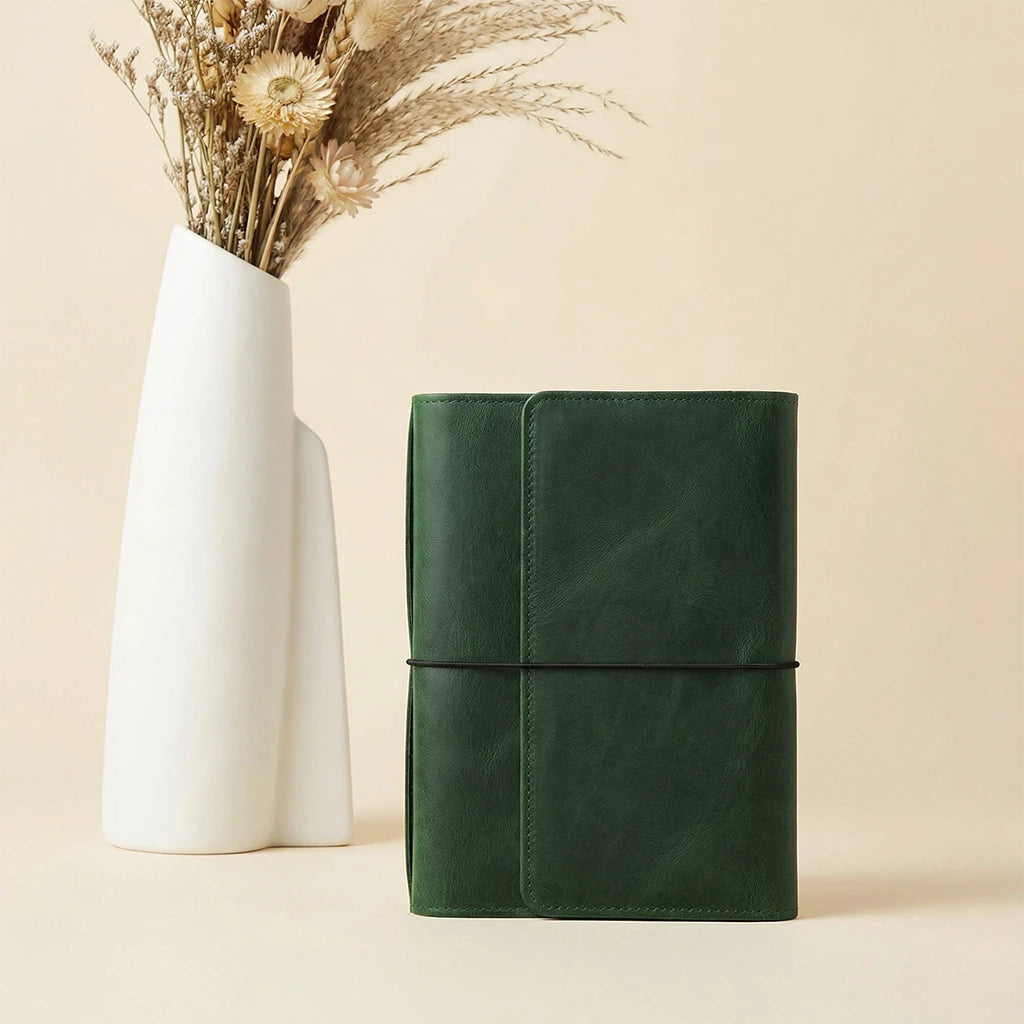 Green leather notebook next to a white vase with dried flowers on a beige background