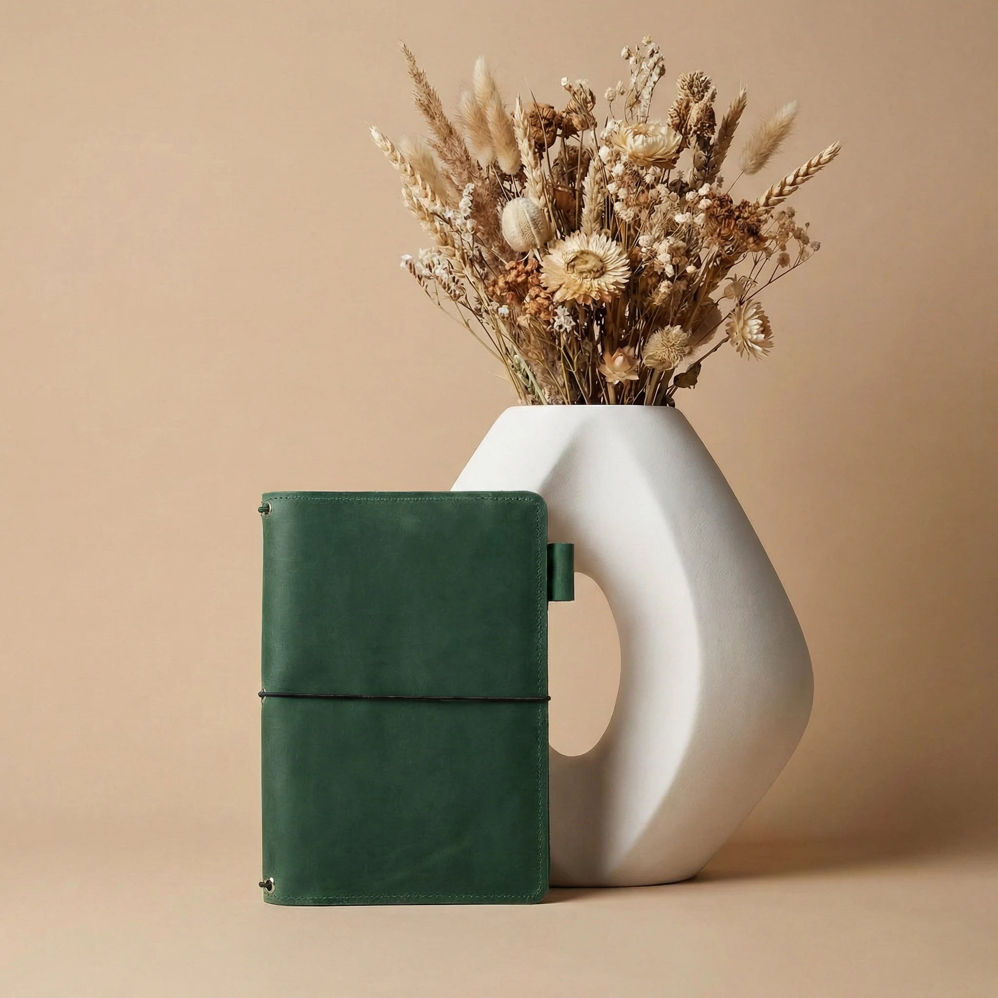 Green leather notebook next to a white ceramic vase with dried flowers on a beige background