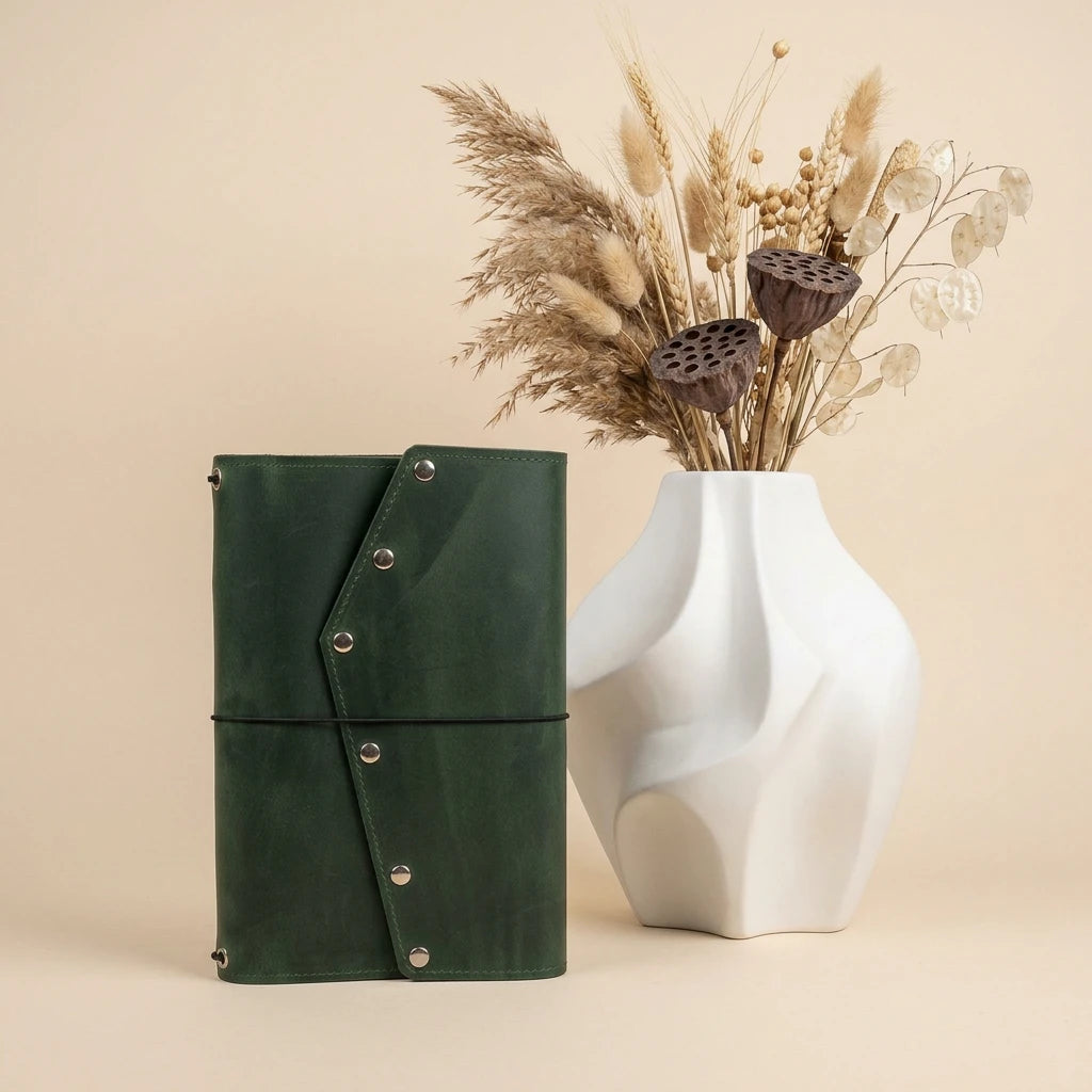 Green leather clutch with silver buttons next to a white vase with dried flowers on a beige background