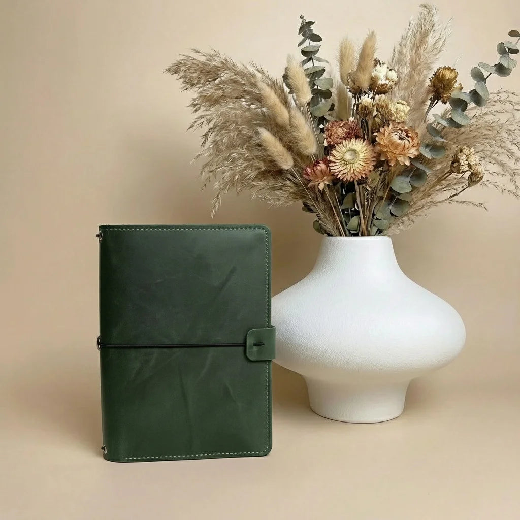Green leather notebook next to a white vase with dried flowers on a beige background