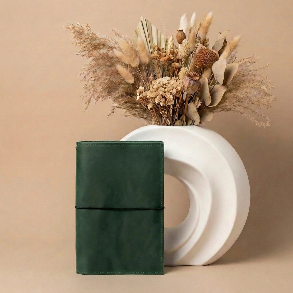 Green leather notebook next to a white vase with dried flowers on a beige background