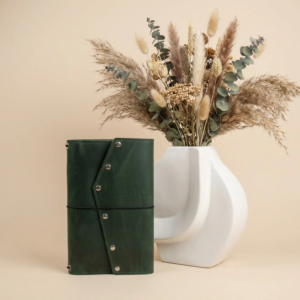 Green leather notebook with silver buttons next to a white vase with dried flowers on a beige background