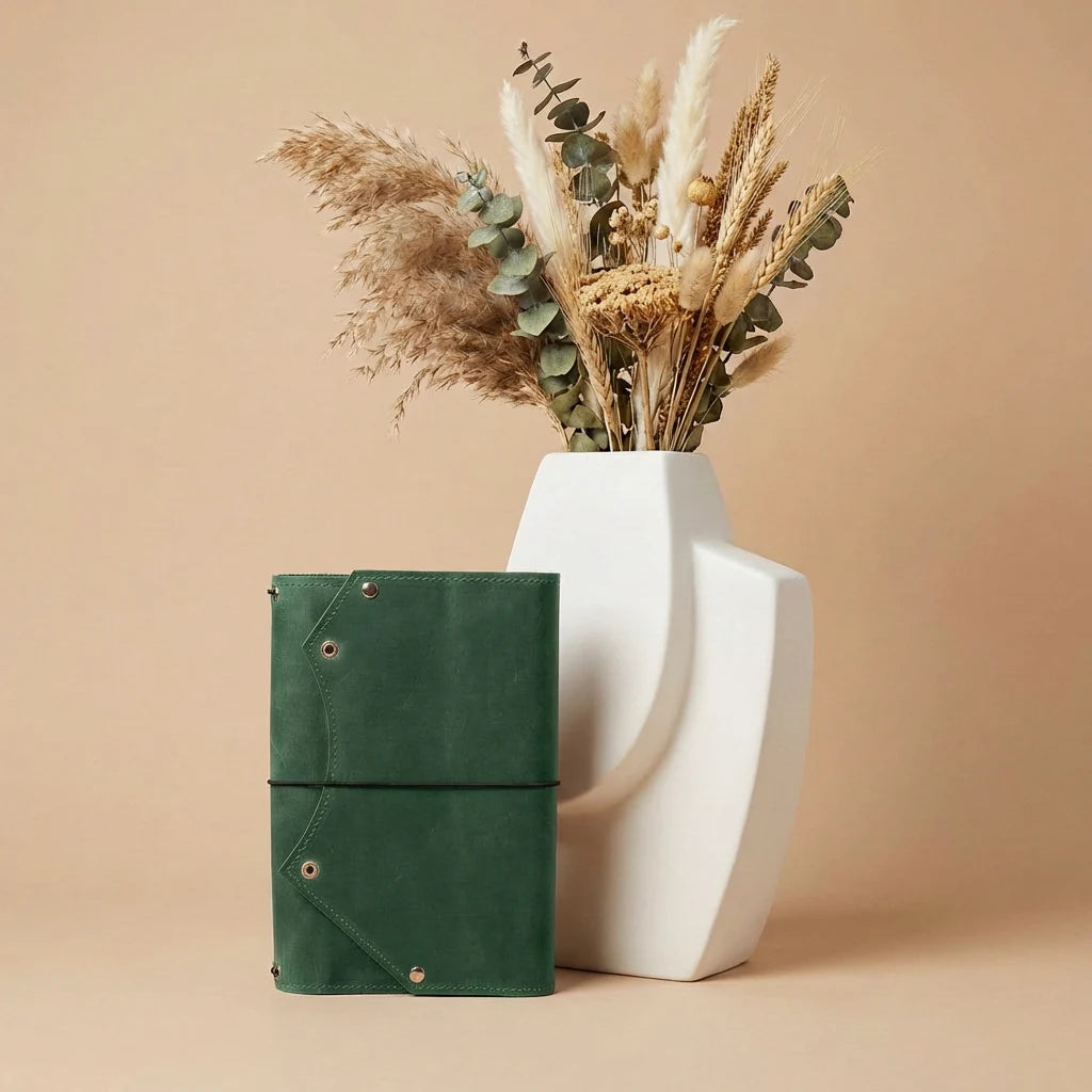 Green leather notebook next to a white vase with dried plants on a beige background