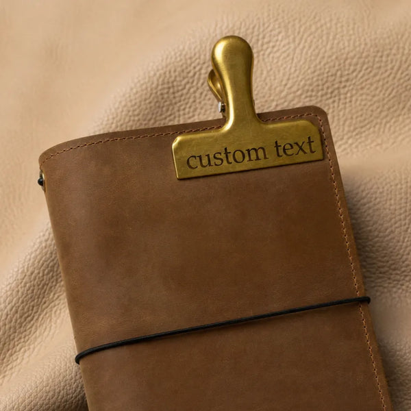 Personalized gold bookmark clip with custom engraving attached to a leather journal cover.