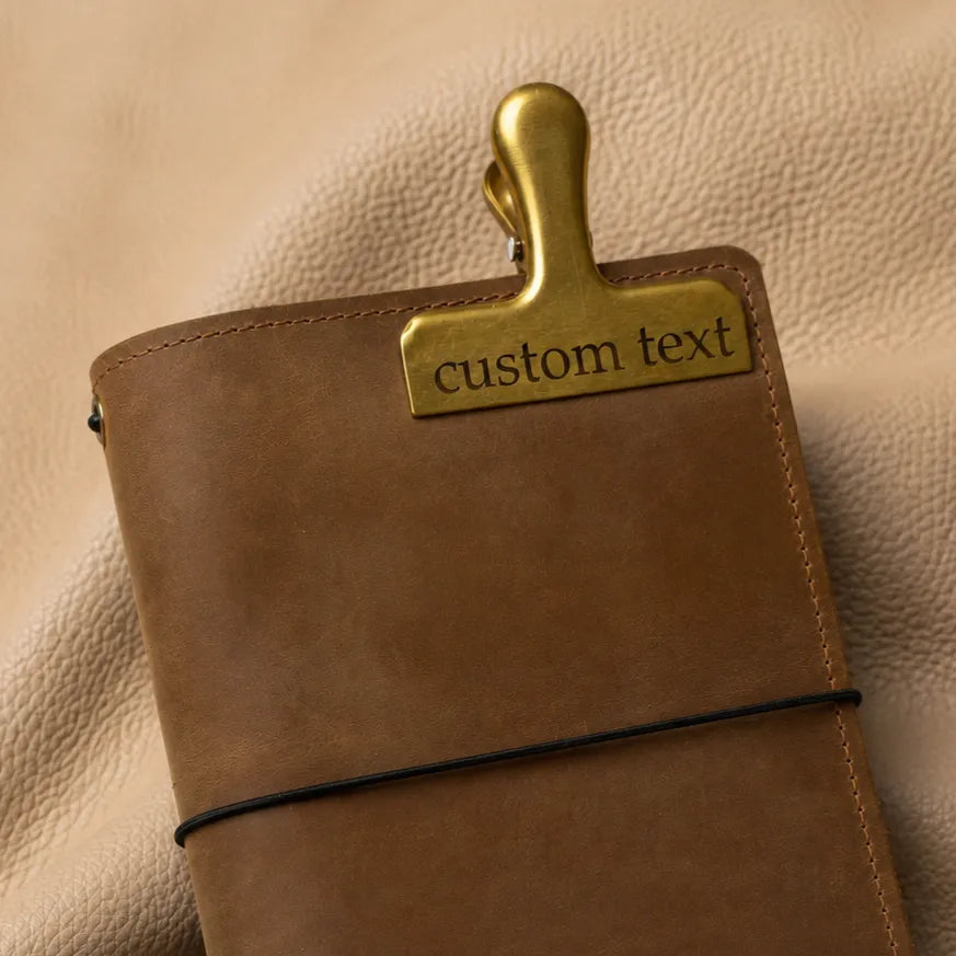 Personalized gold bookmark clip with custom engraving attached to a leather journal cover.