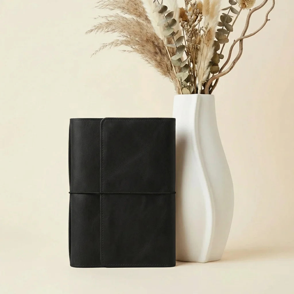Black leather-bound book and white ceramic vase with dried plants on a beige background