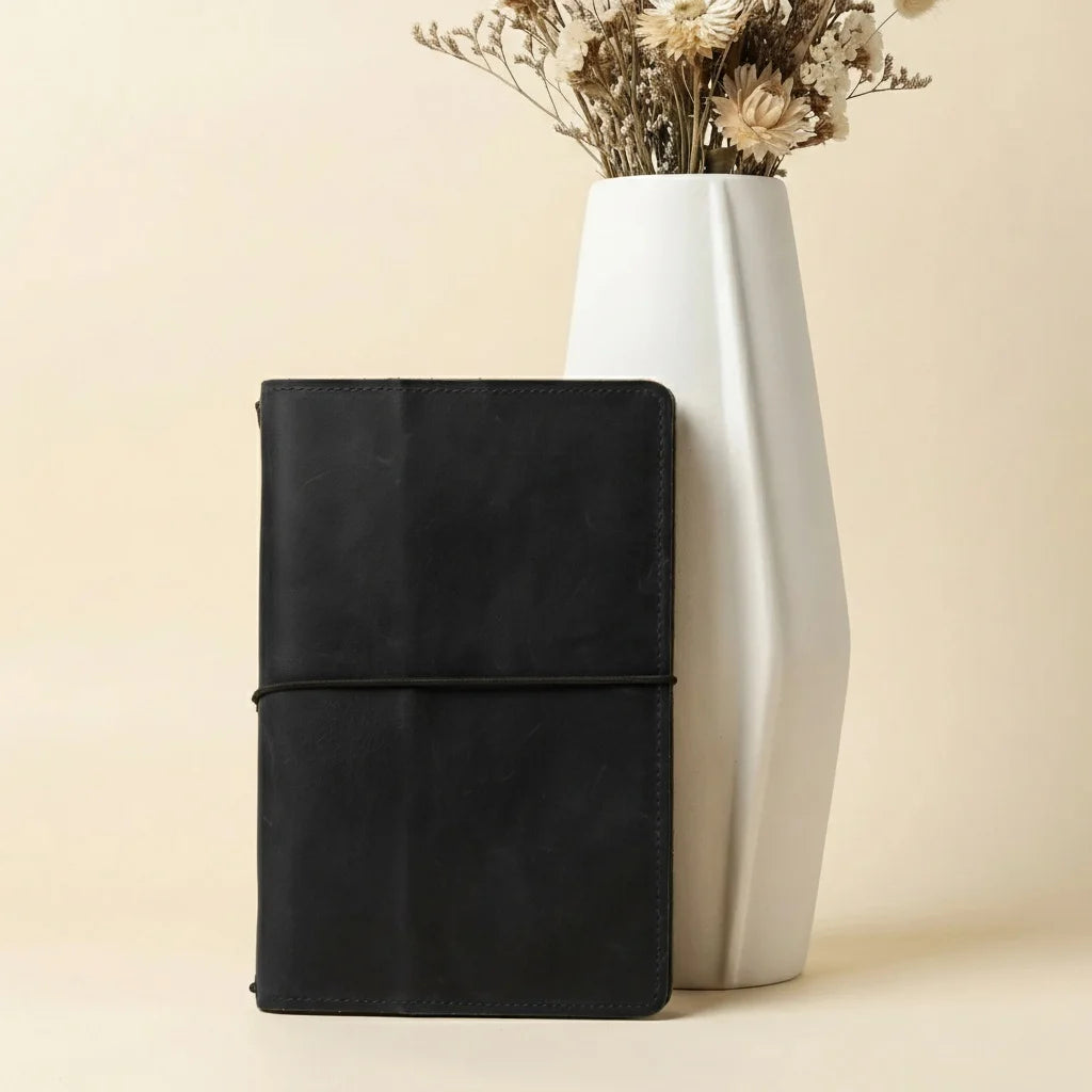 Black leather-bound book and white vase with dried flowers on a beige background