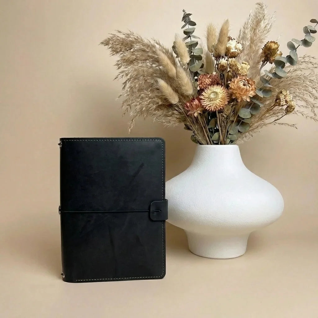 Black notebook next to a white vase with dried flowers on a beige background