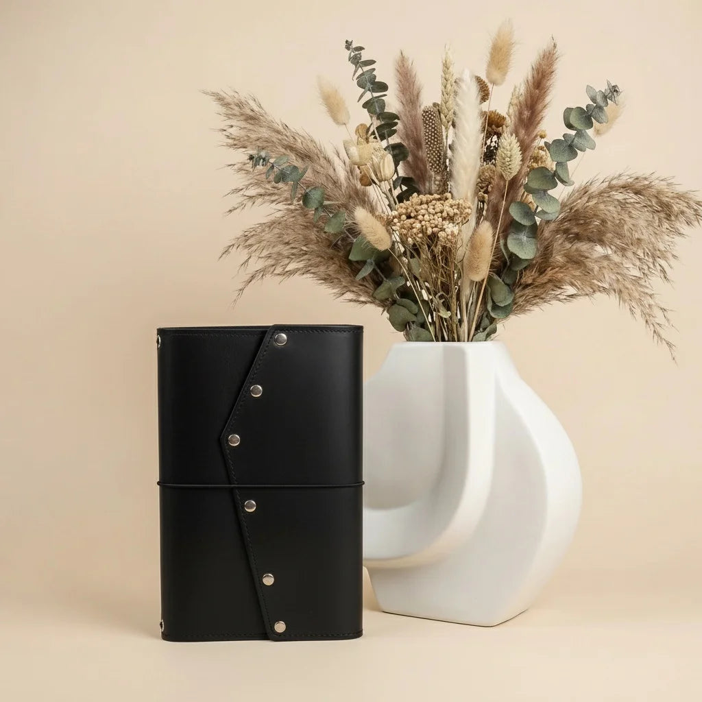Black leather notebook with silver buttons next to a white vase with dried plants on a beige background
