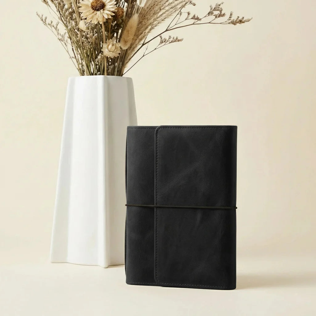 Black leather-bound notebook and white ceramic vase with dried flowers on a beige background