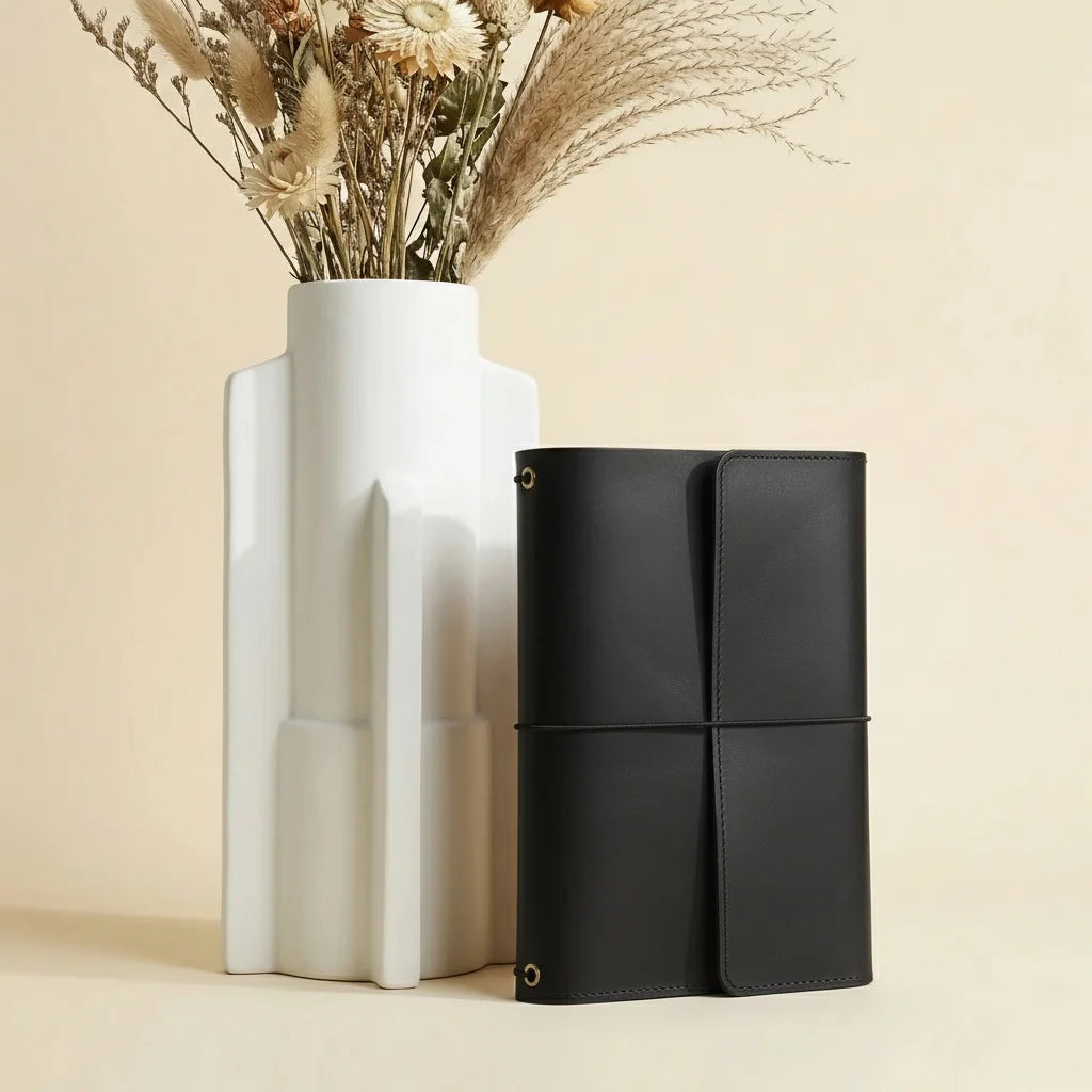 Black leather journal next to a white vase with dried flowers on a beige background