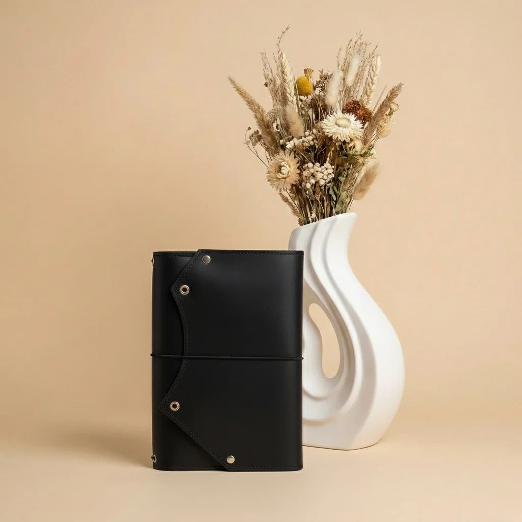 Black leather journal next to a white vase with dried flowers on a beige background