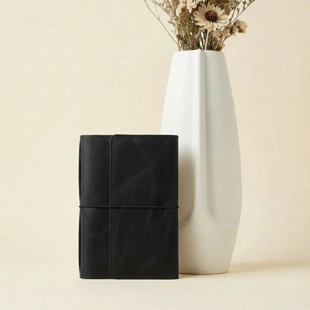 Black leather-bound book and white vase with dried flowers on a beige background