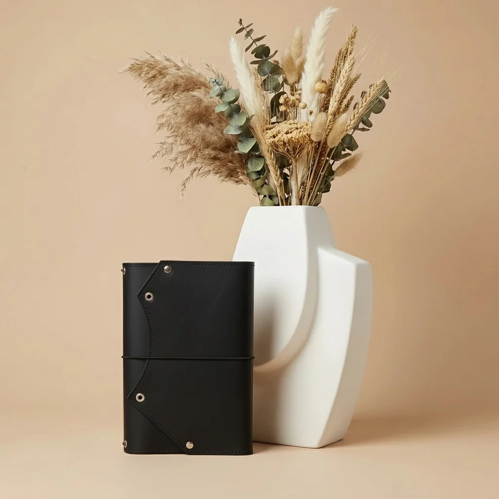 Black leather notebook and white vase with dried plants on a beige background
