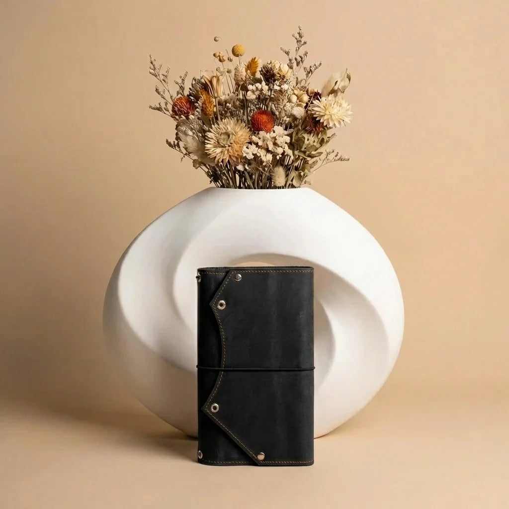 Black leather notebook with gold accents on a white vase with dried flowers against a beige background
