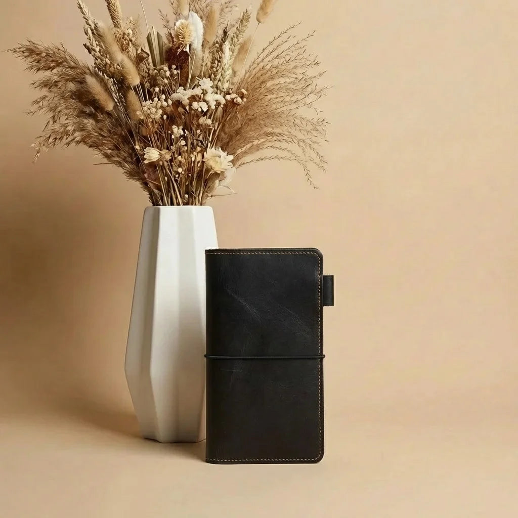 Black leather-bound book with silver buttons next to a white vase with dried plants on a beige background