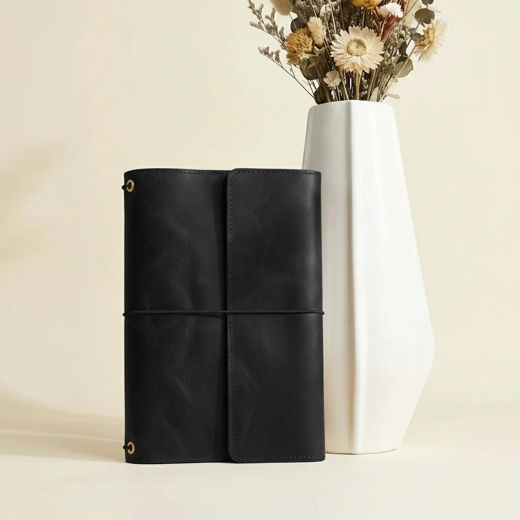 Black leather journal next to a white vase with dried flowers on a beige background