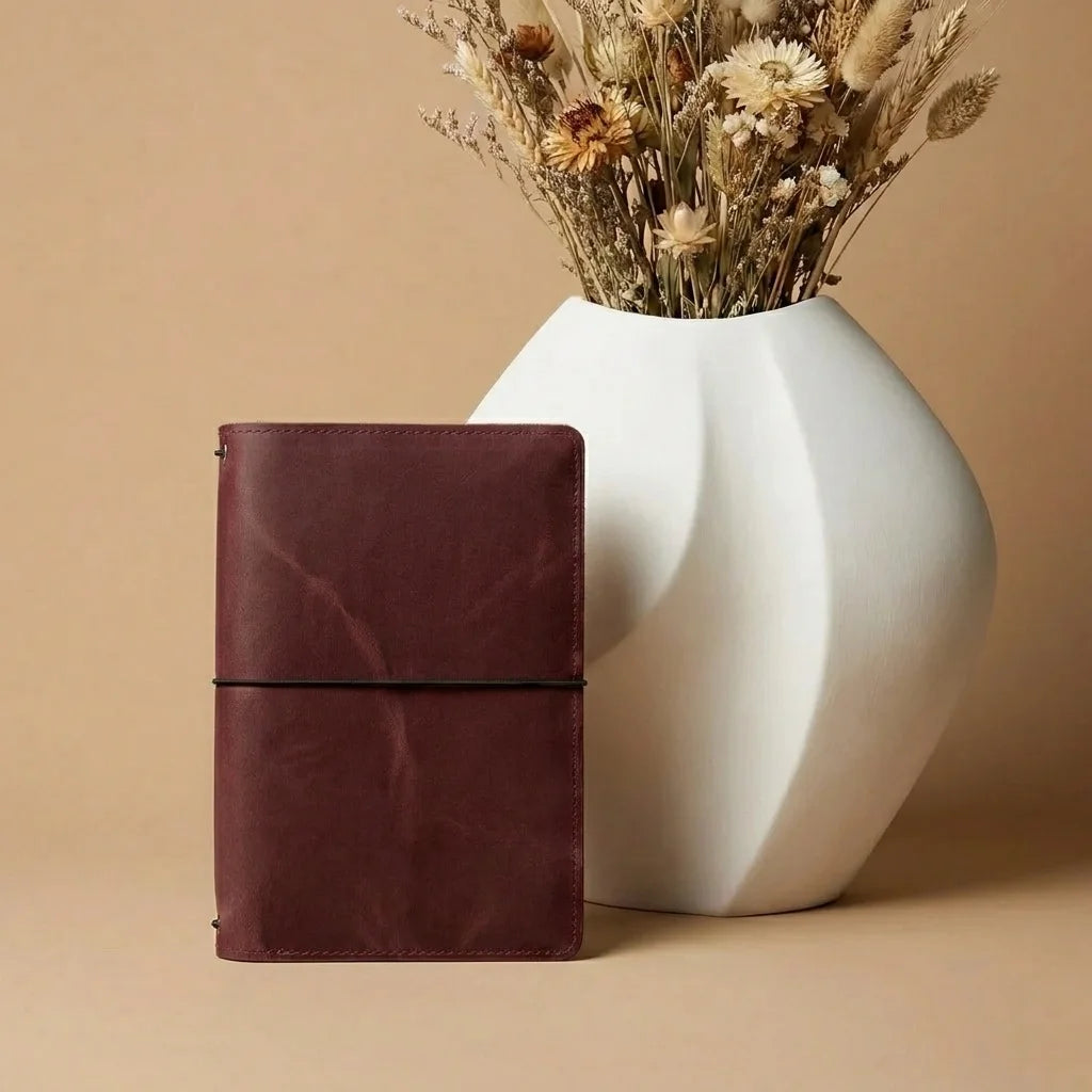 Brown leather journal next to a white vase with dried flowers on a beige background