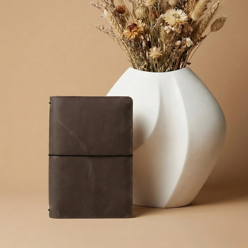 Brown leather notebook next to a white vase with dried flowers on a beige background