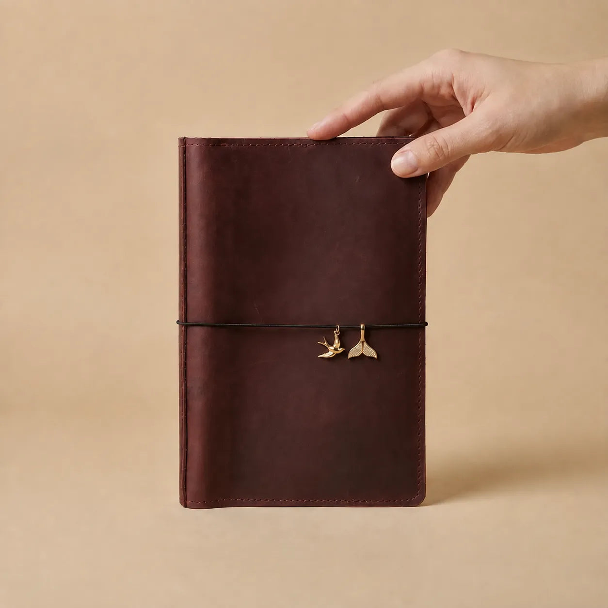 Hand holding a burgundy leather journal with five delicate gold charms on elastic band.